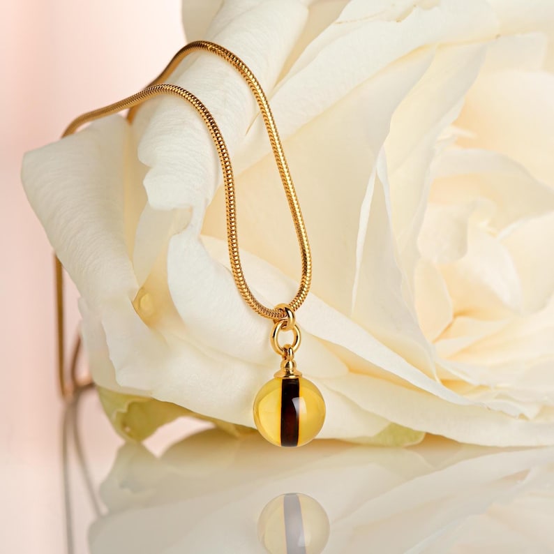 May include: A gold necklace with a yellow pendant and a dark brown stripe. The necklace is set against a white rose. The pendant is reflected on a shiny surface below.