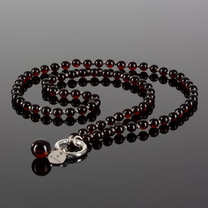 May include: A long necklace featuring dark red, round beads strung together with silver-colored metal. The necklace has a large, matching bead at the end, along with a silver clasp and a small, round tag with text.