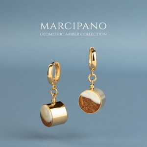 May include: Gold hoop earrings with a dangling geometric amber pendant. The pendant is a cylinder with a white and brown amber design. The earrings are part of the Marcipano Geometric Amber Collection.