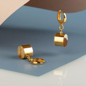 May include: A pair of gold hoop earrings with a dangling cylinder charm. The cylinder is made of gold and has a white and brown resin inlay.