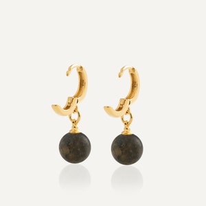 May include: A pair of gold hoop earrings featuring a hinged closure and a dangling dark gray spherical bead. The earrings have a polished metallic finish and a smooth, rounded design, with each bead suspended from a small gold ring at the base.