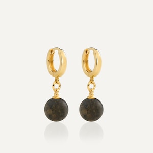 May include: A pair of gold hoop earrings featuring a polished circular band and a dangling dark gray spherical charm. The earrings are connected by a small gold jump ring, showcasing a smooth metallic finish and a matte, textured stone-like sphere.