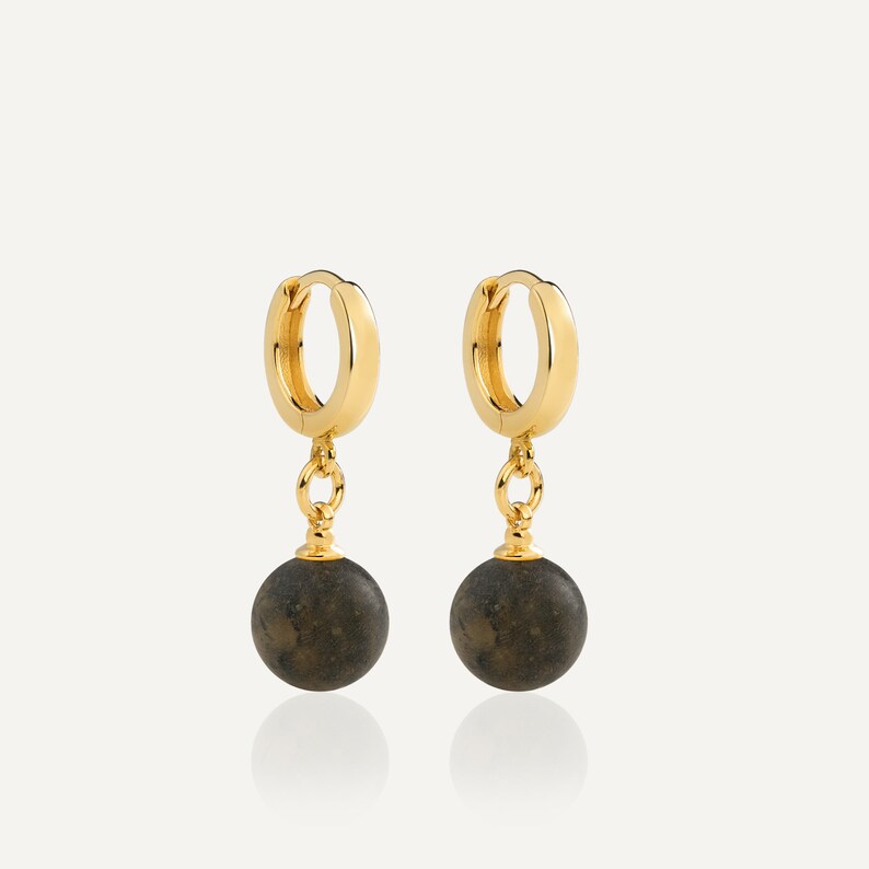 May include: A pair of gold hoop earrings featuring a polished circular band and a dangling dark gray spherical charm. The earrings are connected by a small gold jump ring, showcasing a smooth metallic finish and a matte, textured stone-like sphere.