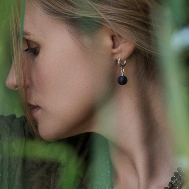 Side profile of a woman framed by soft green foliage, wearing small sterling silver huggie hoop earrings with round black matte Baltic amber drop charms. The natural organic texture of the dark amber beads creates a subtle, elegant contrast. Handcrafted genuine Baltic amber jewelry, ideal as a minimalist daily accessory, unique gift for her, or sophisticated gift for lovers of modern silver gemstone drop earrings.