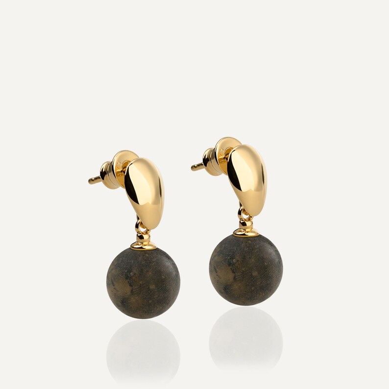 May include: A pair of gold earrings with a dark gray, speckled, round stone pendant. The earrings have a curved, gold bar that connects to the stone.