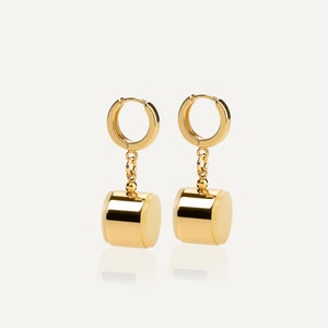 May include: A pair of gold hoop earrings with a cylindrical charm hanging from each hoop. The charms are gold with a white center.