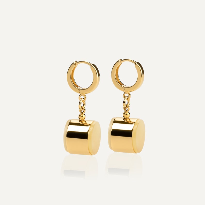 May include: A pair of gold hoop earrings with a cylindrical charm hanging from each hoop. The charms are gold with a white center.