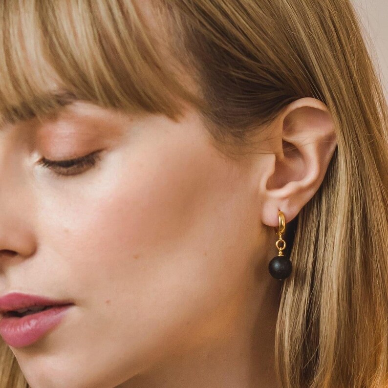 May include: A gold hoop earring featuring a single dangling black spherical bead. The earring is attached to the earlobe, showcasing a polished metallic gold finish on the hoop and a smooth, matte black surface on the round bead component.