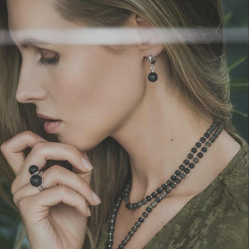 Side profile of a woman framed by soft green foliage, wearing small sterling silver huggie hoop earrings with round black matte Baltic amber drop charms. The natural organic texture of the dark amber beads creates a subtle, elegant contrast.
