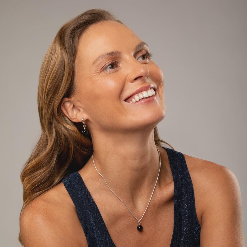 May include: A silver necklace featuring a single round black bead pendant on a thin chain, paired with matching silver drop earrings, each showcasing a small round black bead. The jewelry set is polished and minimalist in its design.