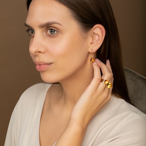 May include: A woman wearing a white shirt with gold hoop earrings and a gold ring with two round amber stones.