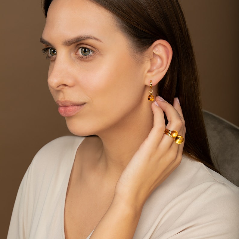 May include: A woman wearing a white shirt with gold hoop earrings and a gold ring with two round amber stones.