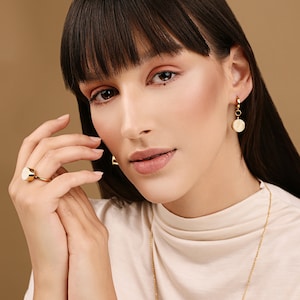 May include: A woman wearing a white turtleneck sweater with a gold chain necklace and gold earrings with a white circular pendant. She is wearing a gold ring with a white circular stone on her finger.