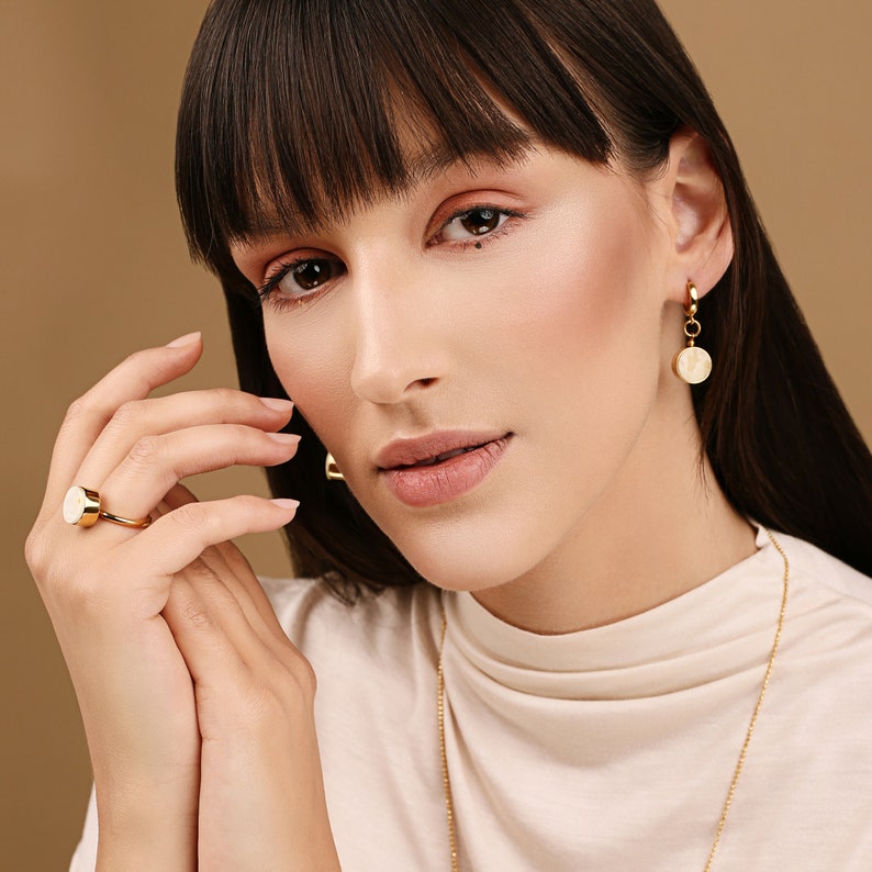 May include: A woman wearing a white turtleneck sweater with a gold chain necklace and gold earrings with a white circular pendant. She is wearing a gold ring with a white circular stone on her finger.