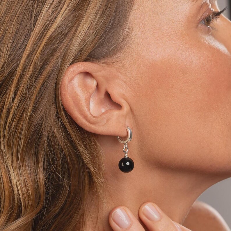 May include: A silver hoop earring featuring a dangling, smooth, spherical black bead. The earring is constructed from a polished silver hoop that connects to a small silver link, which supports the round, glossy black stone or bead component.
