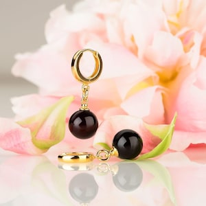 May include: A pair of gold hoop earrings, each featuring a dangling, smooth, spherical black bead. The earrings are crafted with a polished gold finish, showcasing a simple hinged closure mechanism that connects the hoop to the round black charm.