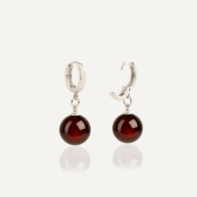May include: A pair of silver hoop earrings, each featuring a dangling, smooth, spherical dark red gemstone. The hoops are polished silver, and the round beads are a deep, translucent garnet color, suspended from a small silver jump ring.