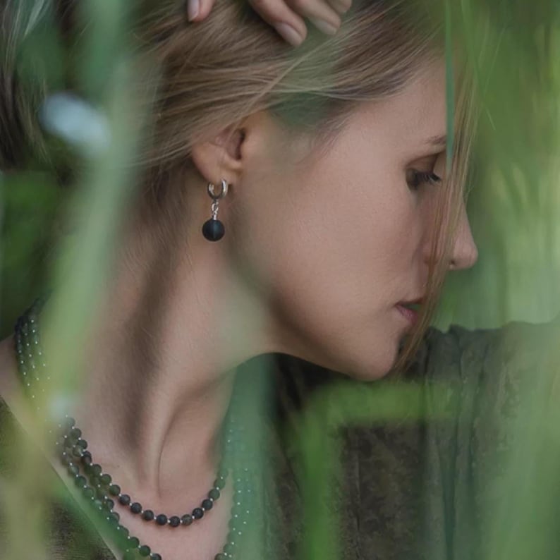 Side profile of a woman framed by soft green foliage, wearing small sterling silver huggie hoop earrings with round black matte Baltic amber drop charms. The natural organic texture of the dark amber beads creates a subtle, elegant contrast.