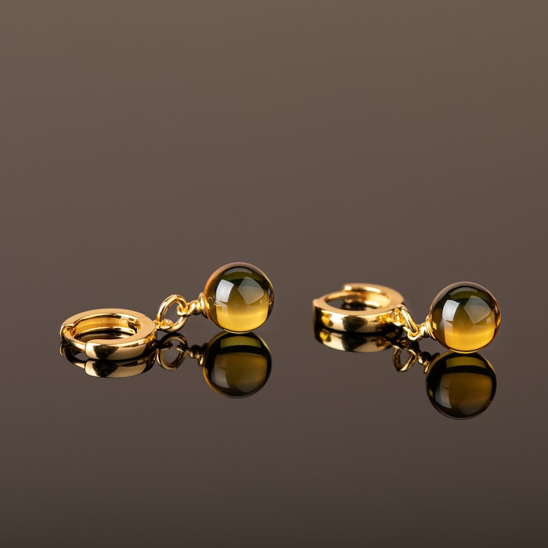 May include: A pair of gold hoop earrings with a dangling green and gold gemstone. The earrings are on a brown surface.