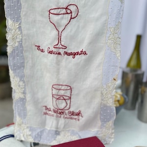Op de afbeelding: Witte linnen cocktail servet met geborduurde rode illustraties van een margarita en een old fashioned. De tekst luidt "The Garcia Margarita" en "The Nelson Blush Mezcal Old Fashioned". De servet heeft een kanten rand.