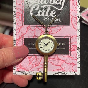 以下が含まれることがあります： 真鍮色の鍵ペンダントネックレス。文字盤には黒い数字と針があり、白い背景です。鍵のペンダントは繊細なチェーンから吊り下げられています。背景はピンクの花柄で、「Quirky Cute Boutique」の文字が入っています。