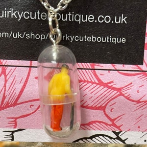 May include: A silver chain necklace with a clear capsule pendant. Inside the capsule is a miniature figure wearing a yellow top and orange pants. The pendant is attached to a silver chain.