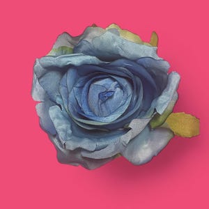 May include: A single artificial rose flower featuring layered petals in shades of blue, ranging from light sky blue to deep navy, with subtle hints of green at the edges of the outer petals, presented against a solid, vibrant pink background.