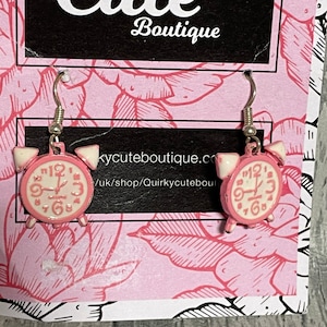 Op de afbeelding: Roze wekker oorbellen met witte wijzerplaten en zwarte wijzers. De oorbellen zijn op een roze en witte bloemenachtergrond met de tekst "Cute Boutique" en een websiteadres.