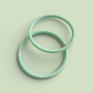 Peut inclure: Deux bracelets circulaires vert clair, entrelacés en forme de huit. Les bracelets ont une finition lisse et brillante, sur un fond vert clair assorti. L'image est une prise de vue minimaliste, de style studio.