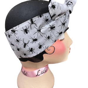 May include: A white headband with a black spider pattern is displayed on a mannequin head. The headband is tied in a bow on top. A pink ribbon around the neck of the mannequin reads "Lulu".