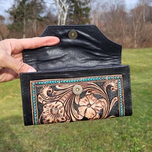 May include: A black leather wallet with a tooled floral design and turquoise accents. The wallet is open and the inside is lined with black leather.