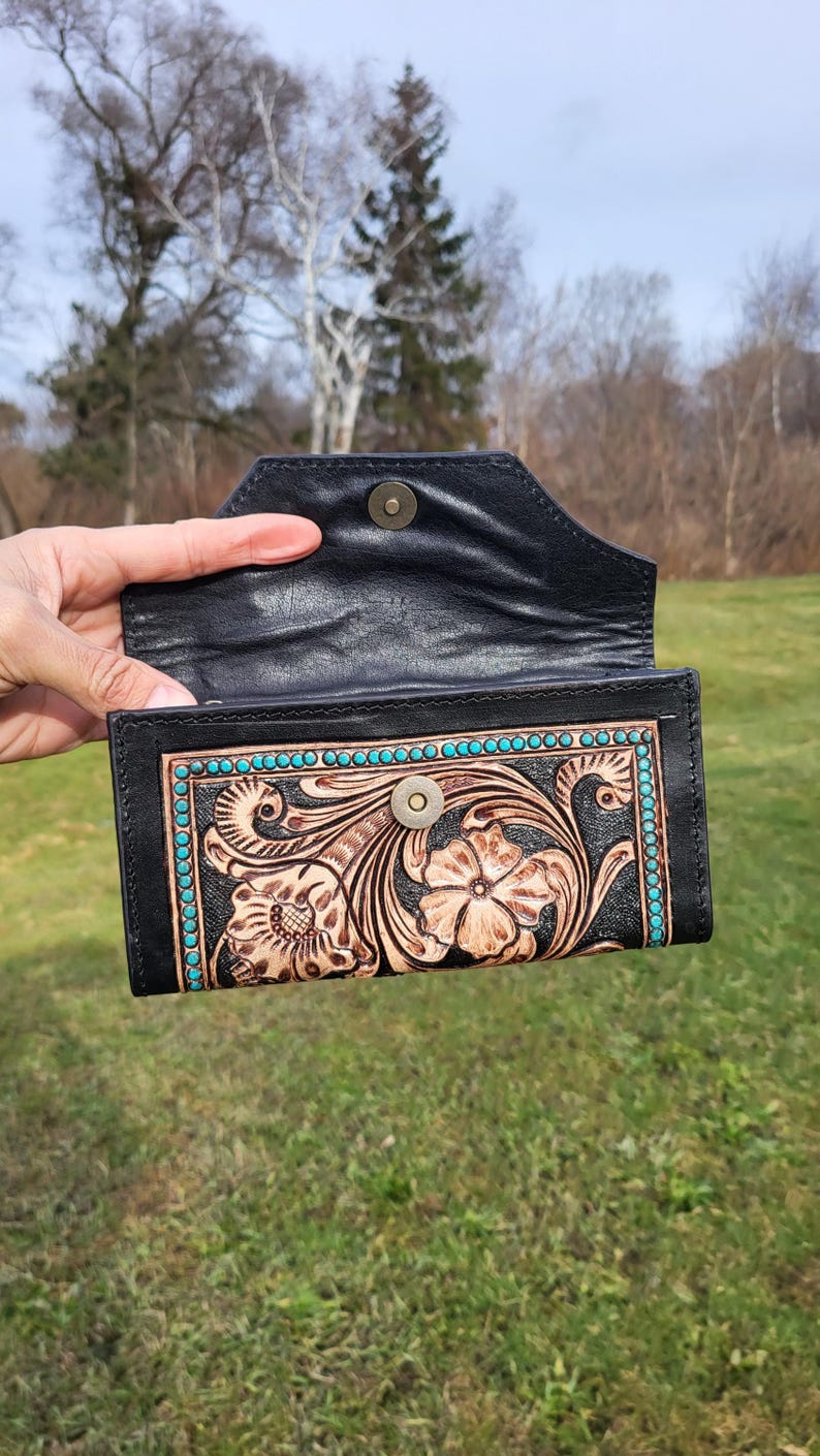 May include: A black leather wallet with a tooled floral design and turquoise accents. The wallet is open and the inside is lined with black leather.