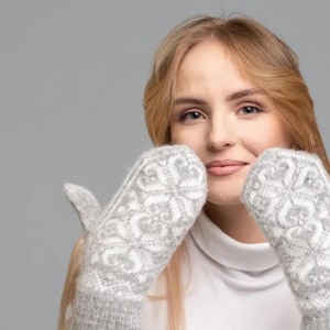 May include: A woman wearing a white turtleneck sweater and gray mittens with a white pattern. The mittens are covering her face.
