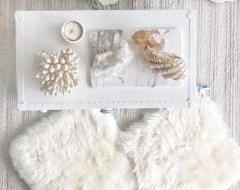 May include: White faux fur cushions on a white wooden table with a white marble serving tray with a white cup, a white coral, a clear crystal, and a beige crystal.