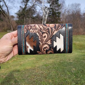 May include: A black leather wallet with a floral design and turquoise accents. The wallet has a white and brown cowhide panel with a geometric pattern.