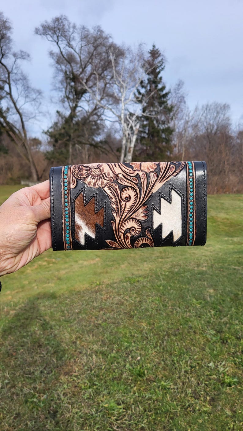 May include: A black leather wallet with a floral design and turquoise accents. The wallet has a white and brown cowhide panel with a geometric pattern.