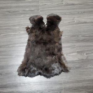 Puede incluir: Una piel de pelaje marrón oscuro, probablemente de un animal, descansa sobre una superficie de madera gris. La piel tiene una apariencia densa y texturizada, con una forma natural. El pelaje es de un color marrón oscuro y rico, con variaciones de tono.