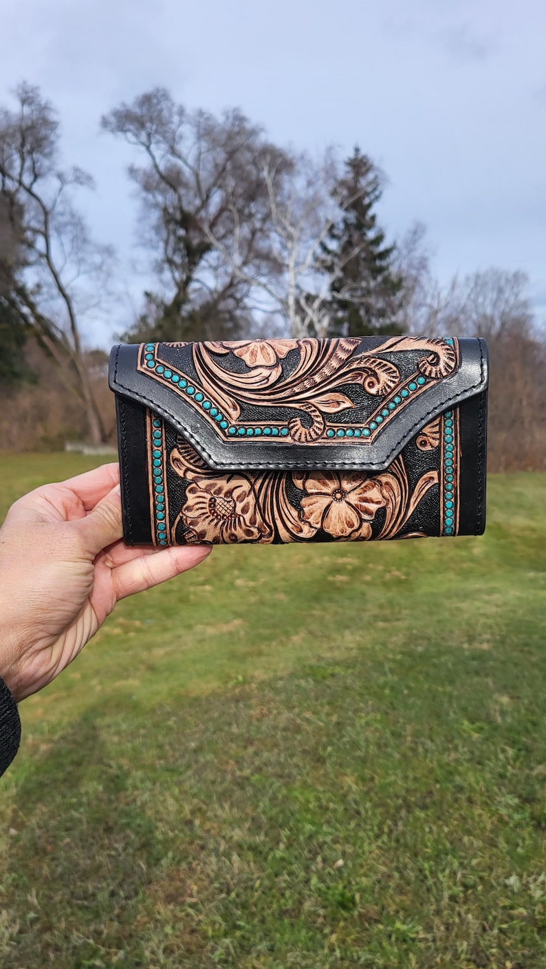 May include: A black leather wallet with a floral tooled design and turquoise accents. The wallet has a flap closure and a decorative stitch detail.
