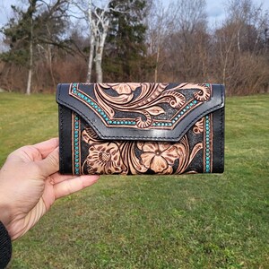 May include: A black leather wallet with a tooled floral design and turquoise accents. The wallet is closed and has a flap.