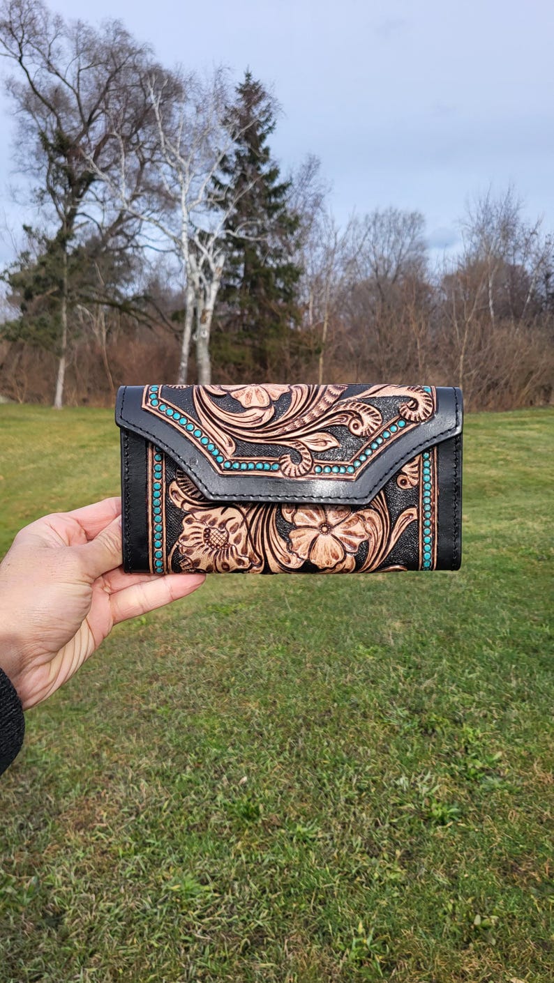 May include: A black leather wallet with a tooled floral design and turquoise accents. The wallet is closed and has a flap.