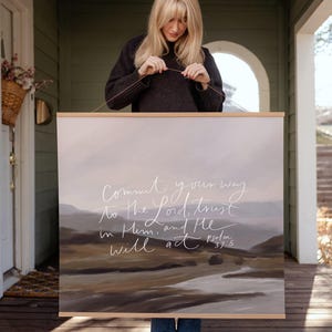 May include: A square wall hanging featuring a landscape painting in muted brown, gray, and white tones. The text "Commit your way to the Lord; trust in Him, and He will act. Psalm 37:5" is written in white cursive script across the center.
