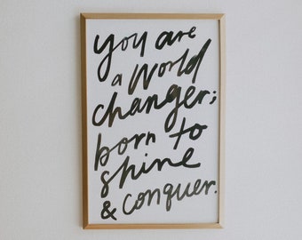 May include: A framed print with a black and white inspirational quote that reads "You are a world changer; born to shine & conquer."