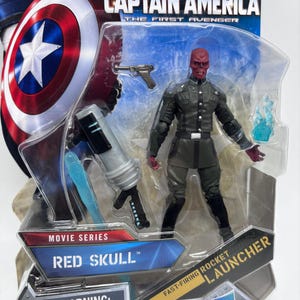 May include: A Red Skull action figure from the Captain America film series, packaged in a clear plastic blister pack. The figure is wearing a dark green uniform and holding a blue energy blast. The packaging includes the text "WARNING: CHOKING HAZARD-Small parts. Not for children under 3 years."