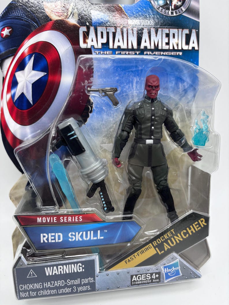 May include: A Red Skull action figure from the Captain America film series, packaged in a clear plastic blister pack. The figure is wearing a dark green uniform and holding a blue energy blast. The packaging includes the text "WARNING: CHOKING HAZARD-Small parts. Not for children under 3 years."