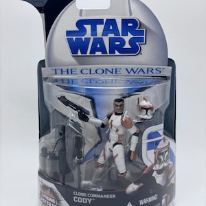 May include: A Star Wars The Clone Wars action figure of Clone Commander Cody in original packaging. The figure is white with orange markings, holding a black blaster rifle, and includes a separate helmet and a firing blaster rifle accessory.