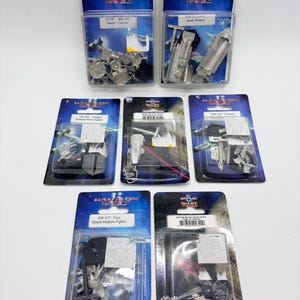 May include: Multiple miniature model kits in clear plastic packaging. The packages feature the "Babylon Wars" logo and product names like "Raider Carrier" and "Gorith Medium Fighter". The models appear to be metallic and are designed for gaming.