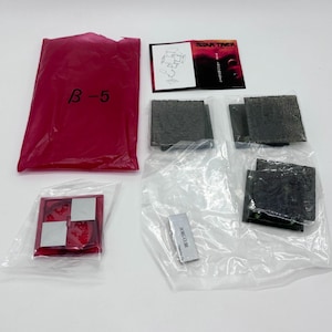 May include: An assortment of Star Trek collectibles, including a red package marked "B-5", several dark gray square pieces, and a small silver "Borg Cube" item. A small instruction sheet and a red and white square are also visible.