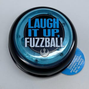May include: A black and blue round toy with the text "LAUGH IT UP, FUZZBALL" in white and blue. The toy has a blue center and a small blue tag with text. The toy is a Star Wars themed item.