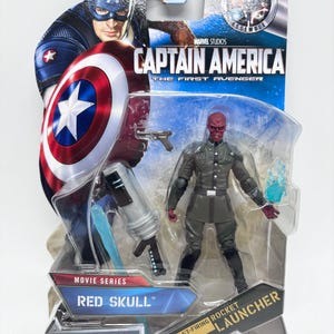 May include: A packaged Red Skull action figure from the Marvel Studios movie series, Captain America: The First Avenger. The figure is in a grey uniform with a red skull face, and comes with a fast-firing rocket launcher. The packaging features Captain America.