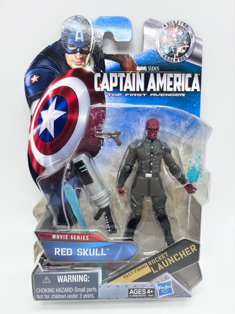 May include: A packaged Red Skull action figure from the Marvel Studios movie series, Captain America: The First Avenger. The figure is in a grey uniform with a red skull face, and comes with a fast-firing rocket launcher. The packaging features Captain America.
